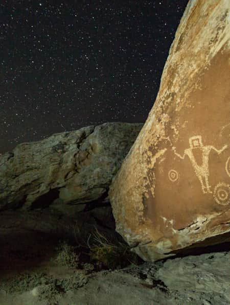 Petroglyphs on a large rock illuminated by soft light, set against a night sky filled with bright stars.