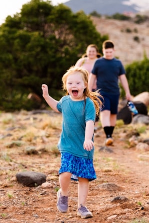 A young girl running on a hiking trail, smiling with her hand up in the air, while her family walks behind her.