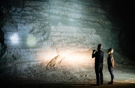 A man and a woman wearing hard hats and holding flashlights, standing next to each other at night, looking at a tall rock slab with various formations.