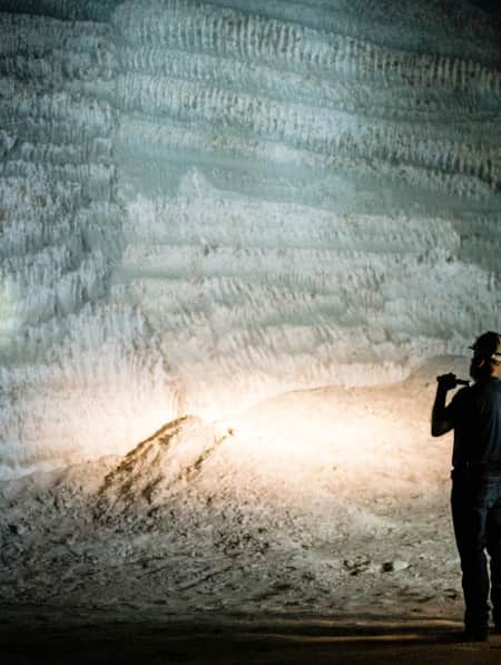 A man and a woman wearing hard hats and holding flashlights, standing next to each other at night, looking at a tall rock slab with various formations.