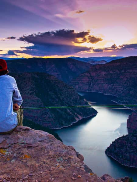 A person holding a fishing rod, overlooking a canyon with a river at the base.