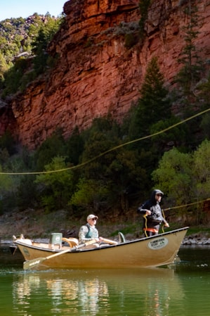 Two people in a small fishing boat, fly fishing off the side at the base of a red mountain.