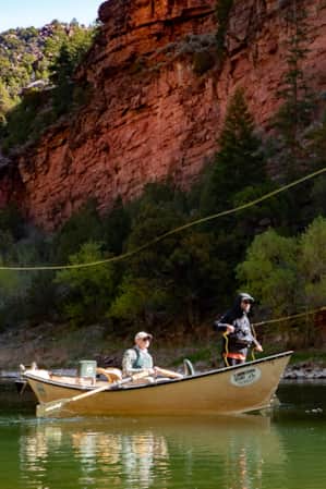 Two people in a small fishing boat, fly fishing off the side at the base of a red mountain.