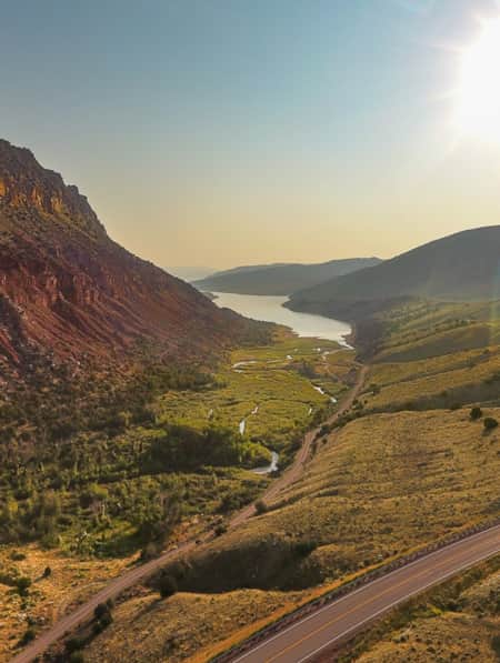 A large red rocky and sandy mountain stands on the left side of the landscape, with a road winding through a lush green valley. A wide stream flows into a narrow river at the mountain’s base.