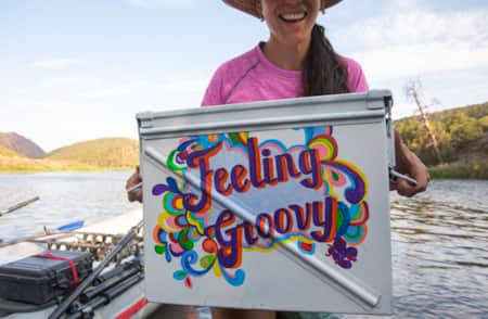 A woman smiling and holding a handwritten sign that reads 'feeling groovy,' with a raft visible behind her at a lake.