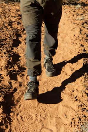 A person's legs walking along a sandy trail with sand on each side covered by a thin layer of a black natural substance.