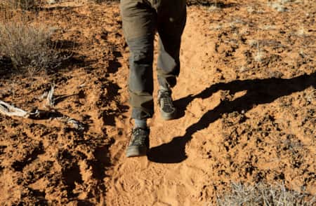 A person's legs walking along a sandy trail with sand on each side covered by a thin layer of a black natural substance.