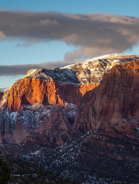 Large red rock mountains with a light dusting of snow, and their peaks illuminated by a ray of sunshine.