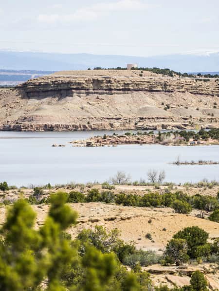 White sandstone rock set upon a hilly landscape, scattered with green bushes, and surrounding a body of water.