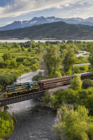 A train crossing a bridge over a stream in a valley, with mountains in the background.