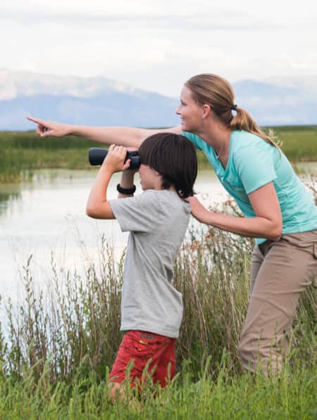 A woman and a young child, standing in the grass by a river, pointing and looking through binoculars at something in the distance.