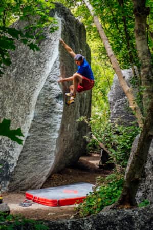 A man climbing a boulder in a forest, surrounded by trees, with a crash pad placed beneath him for a safe landing.