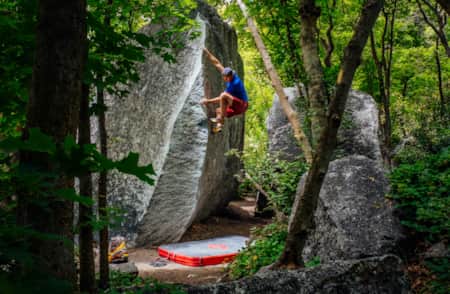 A man climbing a boulder in a forest, surrounded by trees, with a crash pad placed beneath him for a safe landing.