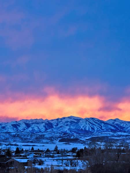 A range of snow-capped mountains set behind a spacious neighborhood at sunset, with deep oranges and pinks filling the sky.