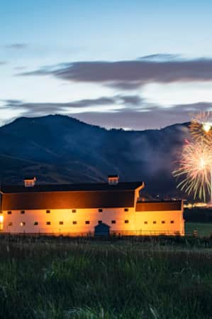 A white barn in a grassy field, illuminated by lights at dusk, set against the silhouette of a mountain range with fireworks bursting to the side.