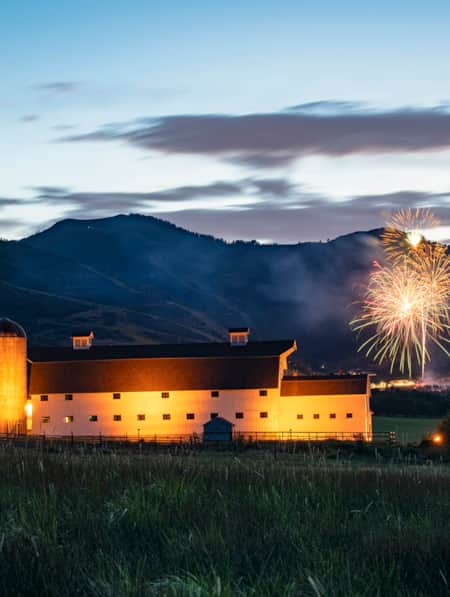A white barn in a grassy field, illuminated by lights at dusk, set against the silhouette of a mountain range with fireworks bursting to the side.