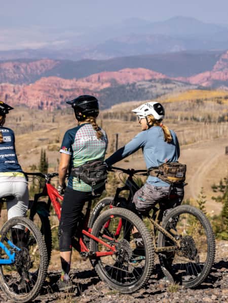 Three women resting on their mountain bikes, overlooking a valley with mountains in the distance.