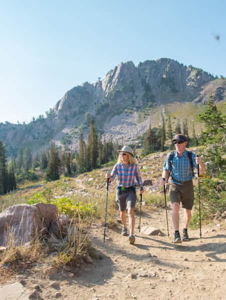 A man and woman hiking with poles on a dirt trail, with a large mountain peak behind them.