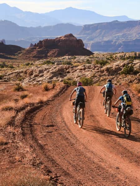 Three people mountain biking on a sandy trail in the desert, heading toward large red rock formations.