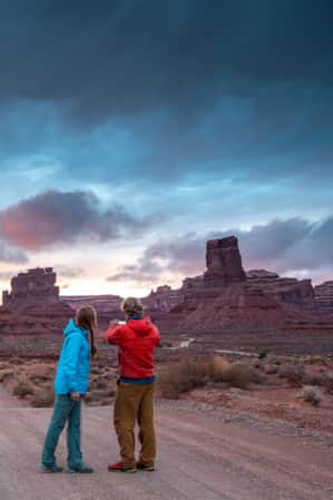 Two people on a gravel road at sunset, with blue hues in the sky, taking a picture of tall, slender rock formations in the distance.