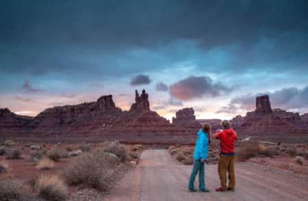 Two people on a gravel road at sunset, with blue hues in the sky, taking a picture of tall, slender rock formations in the distance.