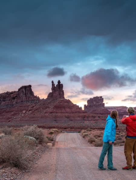 Two people on a gravel road at sunset, with blue hues in the sky, taking a picture of tall, slender rock formations in the distance.