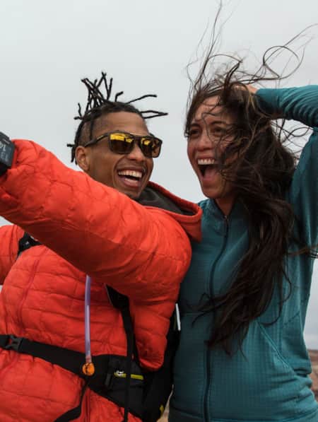 Two people hiking in the desert, smiling at their phone camera for a selfie while the wind blows their hair around.