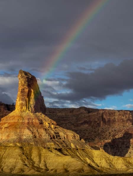 Half of a rainbow arcs over a desert landscape with tall monument rock formations.