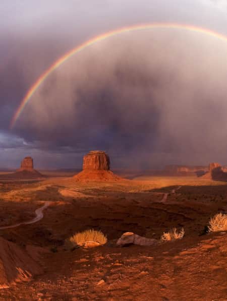Buttes scattered across an open desert field with a full rainbow in the sky.