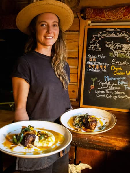 A woman holding two plates of elegantly arranged food in a restaurant with an all-wood interior.