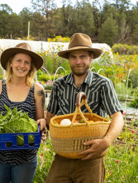 A man and a woman each holding a basket of vegetables, with a garden visible behind them.