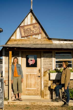 Two women smiling and standing in front of a wooden building on a farm.