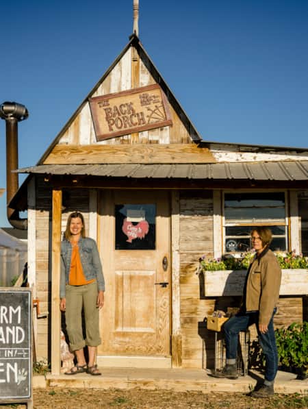 Two women smiling and standing in front of a wooden building on a farm.