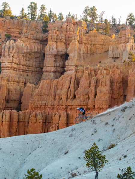 A person on a mountain bike riding down the ridge of a mountain, with hoodoos visible in the distance.