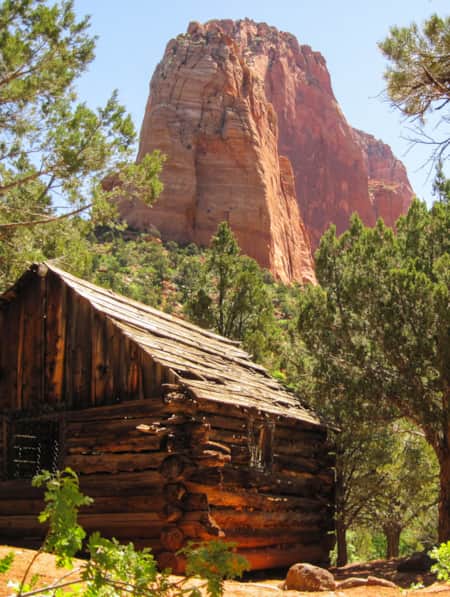 A log house surrounded by trees at the base of a large red rock formation.