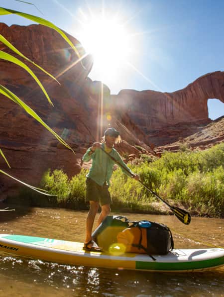 A man stand-up paddleboarding on a river flowing through a red rock canyon during the day.