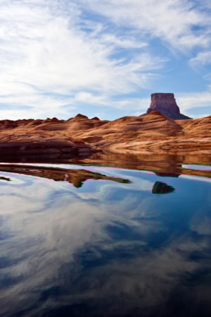 A lake reflecting a blue sky with a few clouds, surrounded by hills of red sandstone.