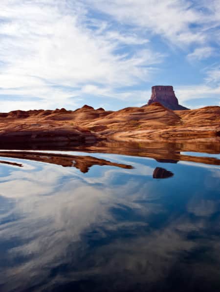 A lake reflecting a blue sky with a few clouds, surrounded by hills of red sandstone.