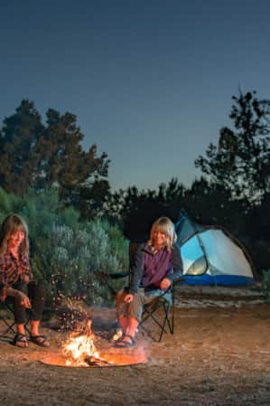 Two women at a campsite with shrubs and their tent behind them, sitting in chairs around a campfire at night, roasting marshmallows.