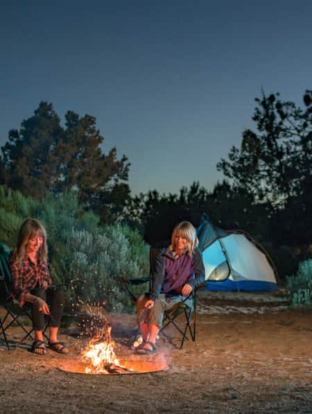 Two women at a campsite with shrubs and their tent behind them, sitting in chairs around a campfire at night, roasting marshmallows.