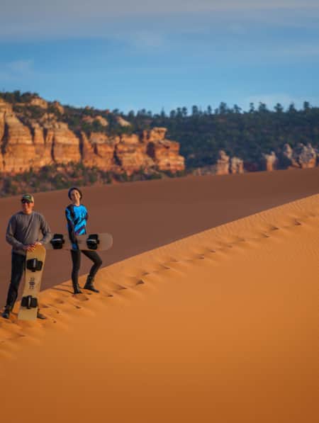 Two people are smiling and holding sandboards to surf down a sand dune at Coral Pink Sand Dunes State Park.