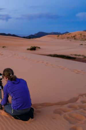 A woman with a tripod capturing photos of sand dunes at dusk."