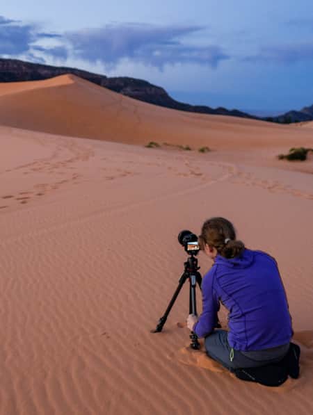 A woman with a tripod capturing photos of sand dunes at dusk."