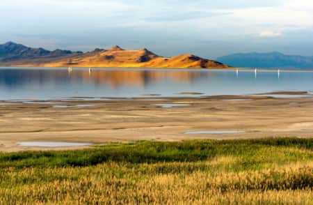 A lake with a sandy shore and green grass beside it, with small mountains visible in the backdrop across the water.
