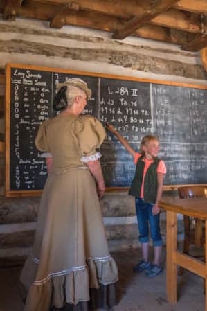 A woman and child in a log homestead. The woman is wearing traditional pioneer clothing, and the young girl is pointing at a chalkboard.