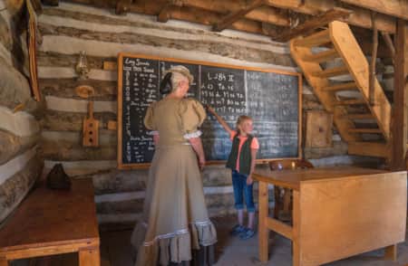 A woman and child in a log homestead. The woman is wearing traditional pioneer clothing, and the young girl is pointing at a chalkboard.
