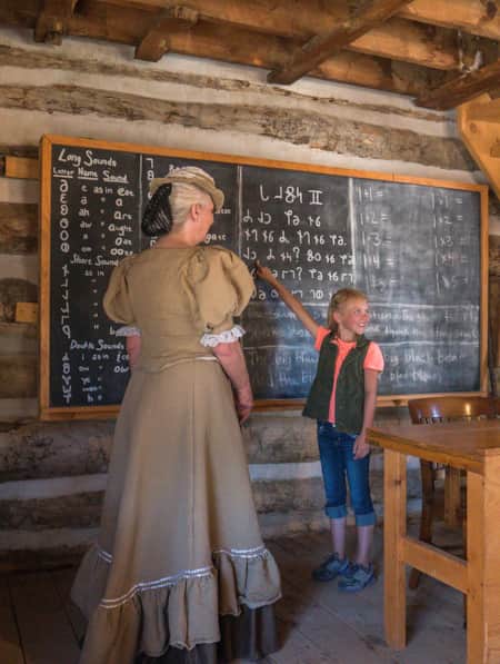 A woman and child in a log homestead. The woman is wearing traditional pioneer clothing, and the young girl is pointing at a chalkboard.