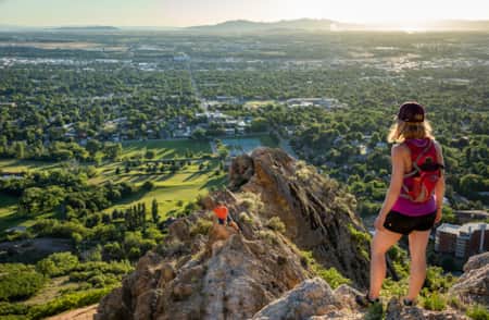 A woman standing on a mountain overlooking a valley with a city below, while a man on a lower rock level looks up at her.