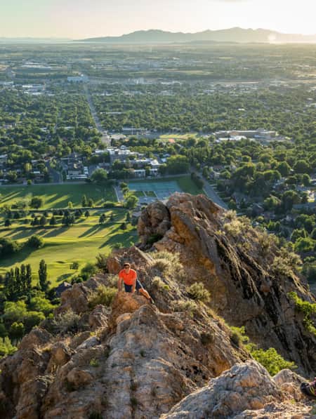 A woman standing on a mountain overlooking a valley with a city below, while a man on a lower rock level looks up at her.