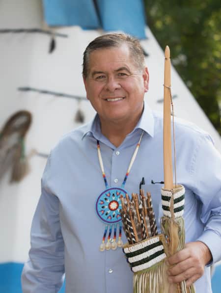 A man wearing a blue button-down shirt stands in front of a white canvas teepee, holding culturally significant artifacts.